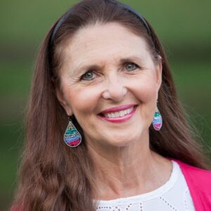 Lynn Eib, author and cancer survivor, smiles while wearing a white blouse and pink sweater and turquoise earrings.